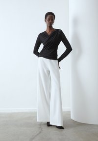 Svart långärmad, omslagsliknande topp tillsammans med högmidjade, vida vita byxor. Bärs med svarta spetsiga skor, mot en minimalistisk bakgrund.