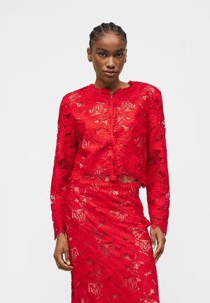 Kvinde iført en rød blomsterblonde bluse med lange ærmer og matchende nederdel, stående mod en ensfarvet lys baggrund.