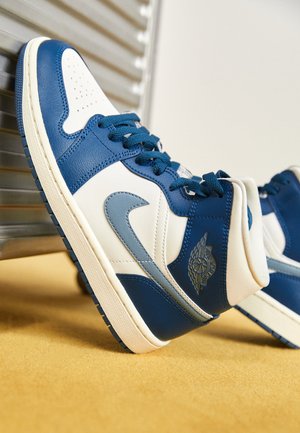 Blå og hvide højt-klædte sneakers med mørkeblå snørebånd og en grå Nike-swoosh, der hviler på en gul overflade nær en metallisk baggrund.