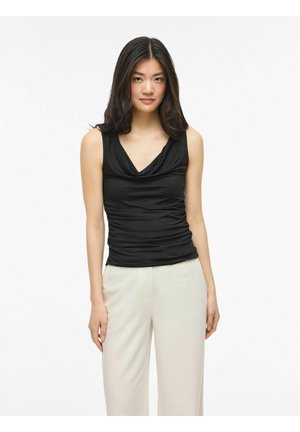 Jeune femme aux longs cheveux foncés porte un haut noir sans manches et un pantalon large beige clair, debout devant un fond blanc uni.