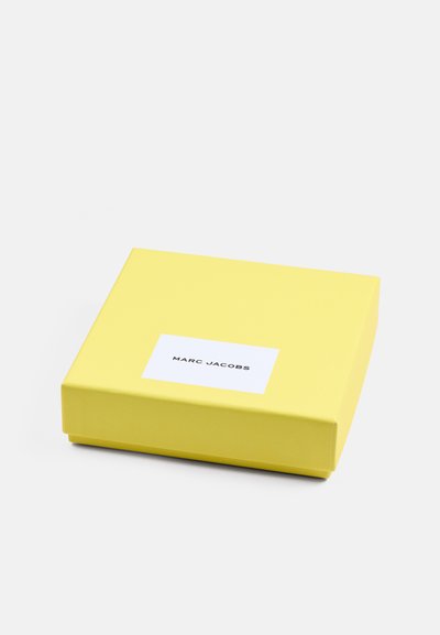 Boîte-cadeau rectangulaire jaune avec une finition lisse, étiquetée d'une étiquette rectangulaire blanche affichant "MARC JACOBS" en police noire.