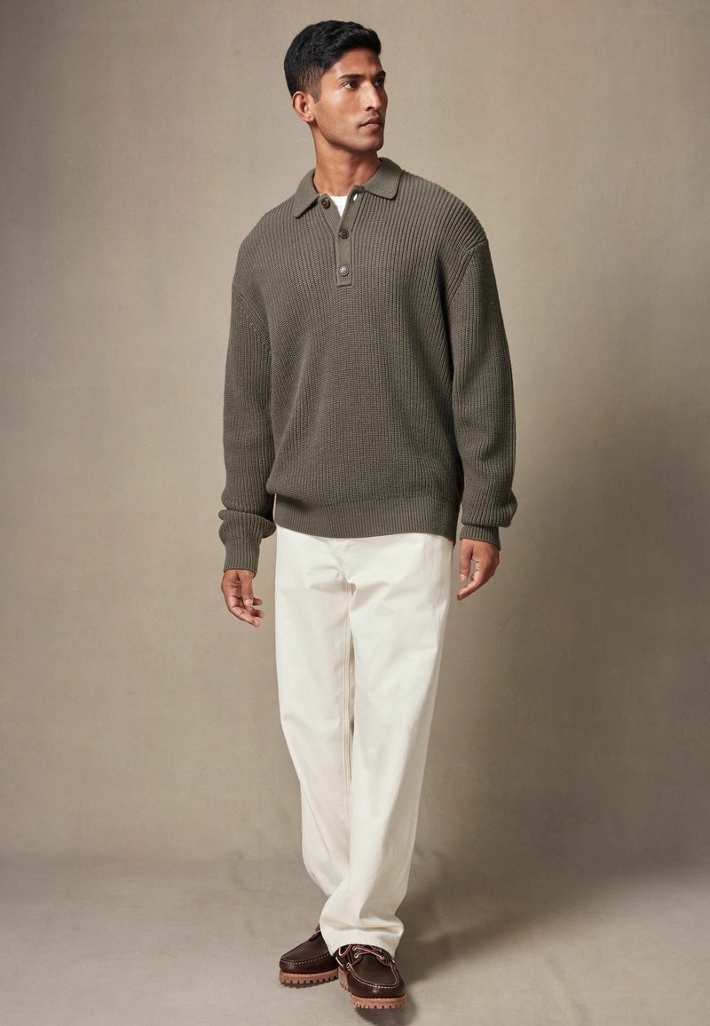 Strikket olivengrønt pullover-sweater med krave og tre knapper, parret med cremede bukser og brune snørebåndssko.