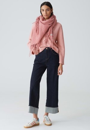 Maglione lavorato a maglia rosa con dettagli di frange, abbinato a jeans scuri a vita alta con orli risvoltati e sneakers multicolori a scarpa bassa.