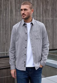 Camisa gris de botones con patrón de puntos blancos, con un bolsillo en el pecho y botones negros, superpuesta sobre una camiseta blanca y jeans oscuros.