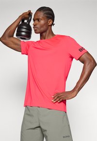 Red athletic shirt with short sleeves featuring a logo on the left sleeve, paired with gray shorts. A black kettlebell is held overhead.