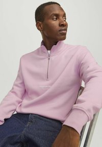 Light pink zip-up sweatshirt with a high collar and embroidered logo on the front, paired with dark blue denim jeans. Smooth texture.