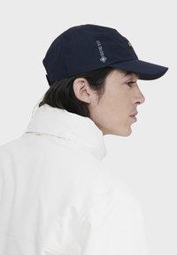 Marineblå cap med en buet skygge, lavet af vandtæt materiale; har et GORE-TEX-logo på siden og bæres sammen med en hvid jakke.