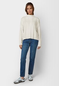 Fleckiger weißer Strickpullover mit hohem Kragen, kombiniert mit blauen Jeans und weißen Sneakern mit schwarzen Streifen und grauen Akzenten.