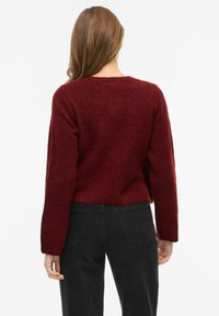 Tamsiai raudonas trumpintas megztinis iš minkštos medžiagos su apvalia iškirpte, ilgomis rankovėmis ir minimaliais siūlais, derinamas su juodais džinsais.