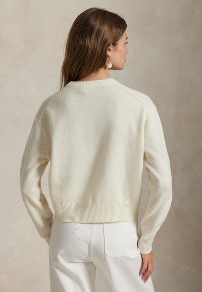 Donna vista da dietro che indossa un maglione color crema e pantaloni bianchi con lunghi capelli castani e orecchini a forma di conchiglia.