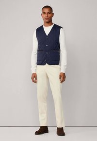 Mörkblå quiltad väst med sex knappar och två framfickor, bärs över en vit tröja, kombinerad med cremefärgade byxor och bruna skor.