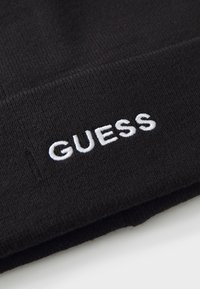 Bonnet en tricot noir avec le texte "GUESS" brodé en blanc. Le tissu présente une texture avec un motif côtelé.