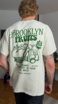 T-shirt de algodão branco com texto verde que diz "BROOKLYN FRUITS" e um gráfico de um caminhão e vegetais, com um corte casual.