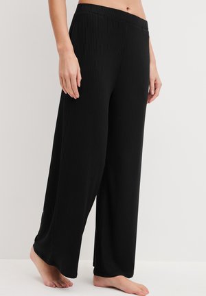 Vrouw die loszittende zwarte geribde brede broek draagt, blootsvoets staand tegen een effen lichte achtergrond.
