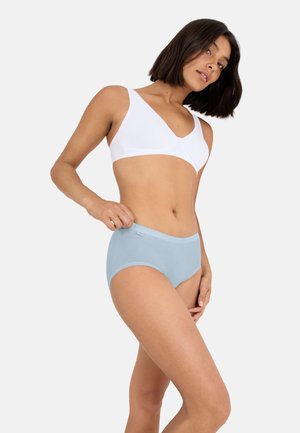 Žena s krátkými tmavými vlasy, oblečená v bílém podprsence a světle modrých kalhotkách s vysokým pasem, pózující na jednobarevném bílém pozadí.