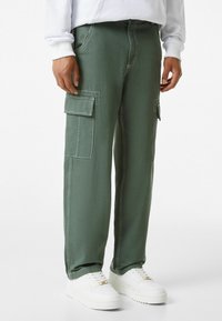 Olivgröna cargobyxor i bomull med kontraststygn, med två sidofickor och en bakficka, kombinerade med vita sneakers.