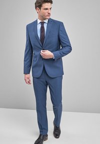 Hombre ajustando un botón de un traje azul a medida con camisa blanca, corbata de estampado y zapatos de vestir negros, de pie contra un fondo gris.