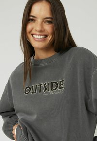 Sweatshirt gris avec un col rond et des manches longues. Caractérisé par l'inscription "OUTSIDE" brodée en noir et blanc, avec le texte "nouvelles horizons" en dessous.