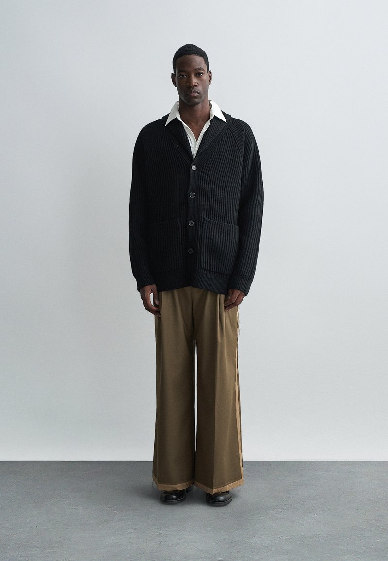 Cardigan nero a coste con bottoni grandi e due tasche, indossato sopra una camicia bianca con colletto, abbinato a pantaloni larghi color tan e scarpe nere.