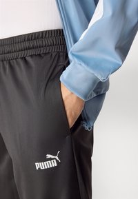 Sorte sportbukser med en glat overflade, udstyret med en elastisk talje, sidelommer og et hvidt Puma-logo. En lyseblå jakkeærme er synlig.