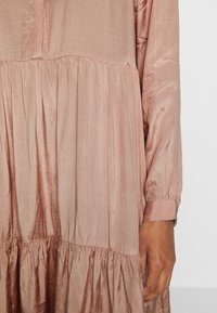 Abito in satin rosa polveroso a maniche lunghe con strati arricciati, maniche con polsini e una mano visibile che riposa di lato.
