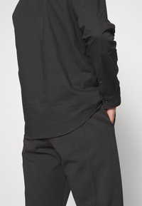 Personne portant une chemise noire à manches longues porté par-dessus un pantalon foncé, avec une main dans la poche, sur un fond clair uni.