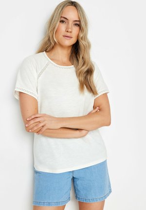 White short-sleeve top with a round neckline and decorative trim along the sleeves. Styled with light blue denim shorts.