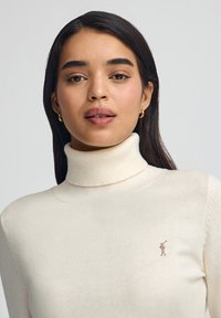 Maglione dolcevita color crema realizzato in tessuto morbido, con un piccolo dettaglio logo sul petto e un design aderente con polsini a coste.