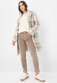 Vrouw in een beige en grijs geruite jas, crèmekleurige sweater, bruine broek en witte sneakers, poserend tegen een effen lichte achtergrond.