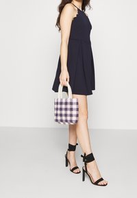 Musta halterneck-mekko, jossa on aallotettu kuvio, yhdistettynä pieneen kudottuun laukkuun violetin ja valkoisen sävyissä sekä mustiin nilkkaremmillä varustettuihin korkokenkiin.