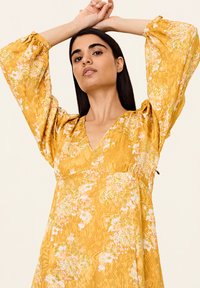 Rochie florală galbenă cu decolteu în V, mâneci lungi fluide, croială lejeră și material texturat cu modele florale roz și verzi.