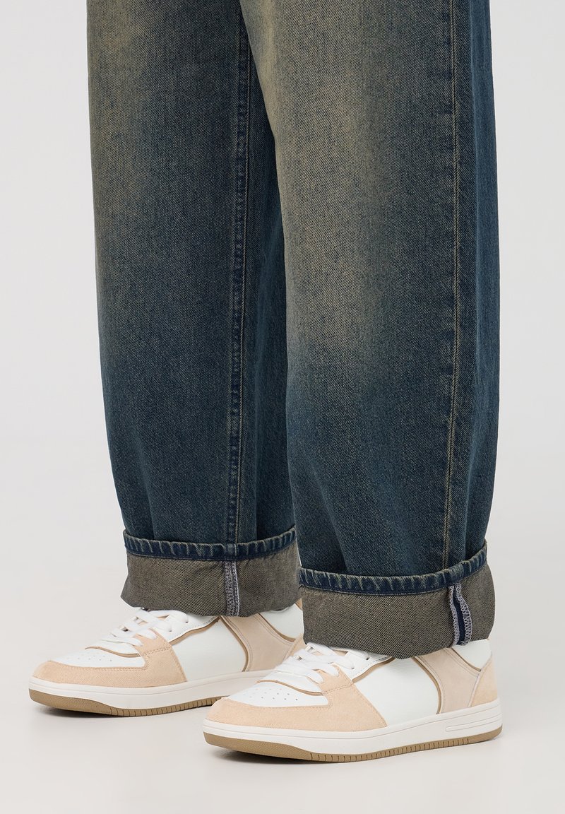 Sneaker bianchi e beige con dettagli in suede e pelle liscia, dotati di punta perforata, lacci piatti e suola in gomma beige, abbinati a jeans in denim scuro.