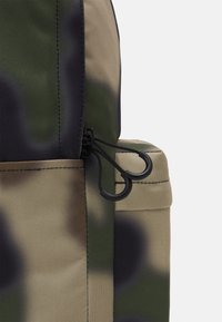 Närbild på en ryggsäck med camouflage-mönster som visar en dragkedja med två svarta ringformade dragknoppar på en sidoficka.