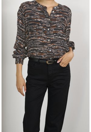 Kvinde iført en langærmet skjorte med knapper og et mønster i sort, brunt og hvidt, stoppet ned i sorte bukser med sort bælte.