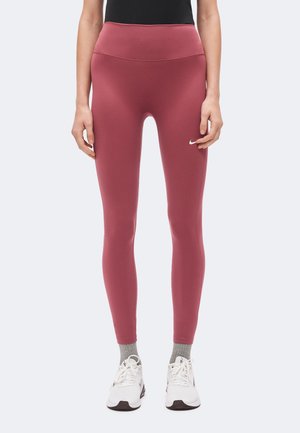 Person iført højtaljede lilla leggings med et lille Nike-logo på låret, grå sokker og hvide Nike-sneakers, der står oprejst.