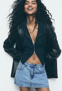 Giubbotto di pelle nera sopra un maglione blu navy con zip e una mini gonna in denim azzurro, con una texture liscia e un design aderente.