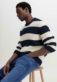 Strickpullover mit marineblauen und cremefarbenen Streifen, Rundhalsausschnitt und gerippten Bündchen, kombiniert mit blauen Jeans. Modell sitzt auf einem Holzstuhl.