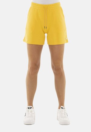 Persoon draagt felgele shorts met trekkoord en witte sneakers, staand tegen een effen lichte achtergrond.