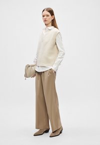 Colete de malha marfim sobre uma camisa branca com colarinho, combinado com calças largas bege e botas de cano curto taupe com salto; segurando uma bolsa com pregas.