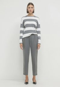 Donna che indossa un maglione a righe bianco e grigio, pantaloni grigi su misura e décolleté neri a punta, in piedi contro uno sfondo bianco semplice.