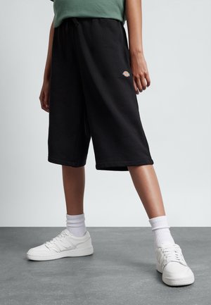 Osoba w czarnych, do kolan szortach z małym logo, białych skarpetkach, białych sneakersach, stojąca na szarej podłodze na tle jednolitego tła.
