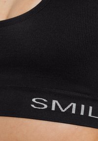 Zwarte geribde bralette met een ronde halslijn en een stoffen band met "SMILE" in het wit gedrukt. Zachte textuur met een minimalistisch ontwerp.