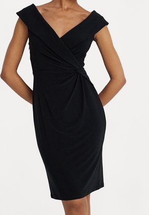 Robe noire sans manches longueur genou avec un décolleté en V épaules dénudées et un détail noué froncé à la taille sur une personne debout.