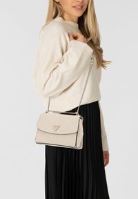 Donna che indossa un maglione crema e una gonna nera plissettata, che porta una borsa a tracolla beige con una tracolla a catena sulla spalla.