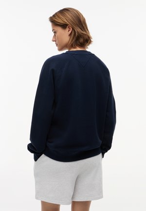 Άτομο με κοντά καστανά μαλλιά, φορούν μπλε σκούρο φούτερ και ανοιχτό γκρι σορτς, στέκεται με τα χέρια στις τσέπες, στραμμένο προς τα αριστερά σε λευκό φόντο.