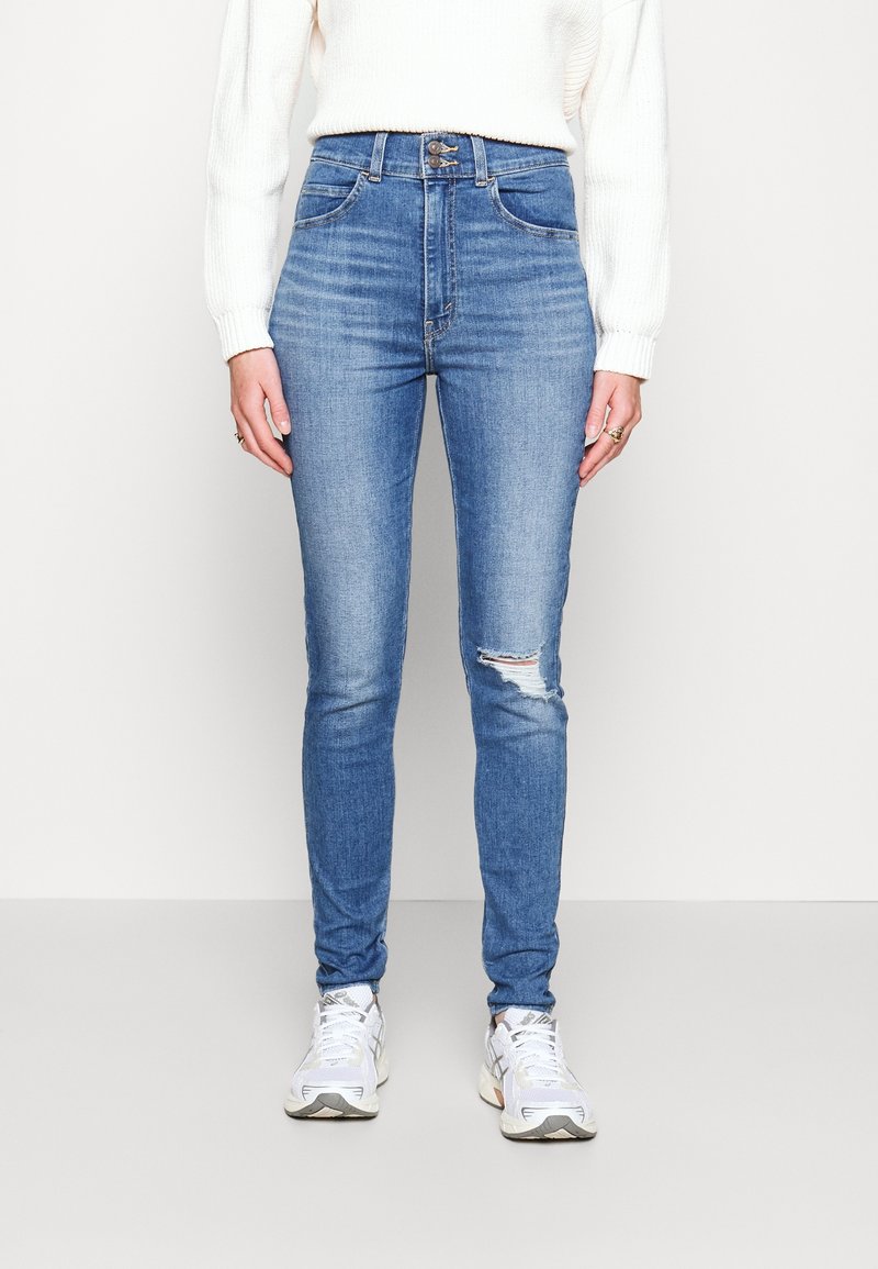 Personne portant un jean skinny bleu taille haute avec une petite déchirure sur un genou, un pull blanc et des baskets blanches, debout devant un fond uni.