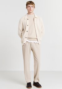 Cardigan lavorato a maglia beige con colletto, chiusura con bottoni, indossato sopra un maglione beige chiaro e pantaloni crema. Scarpe scure completano il look.