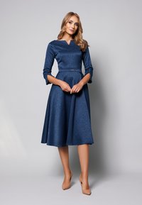 Granatowa sukienka z dopasowaną górą i rozkloszowanym dołem, z teksturowanym wzorem w kropki i rękawami do łokcia. Gładki materiał, subtelny pasek w talii.