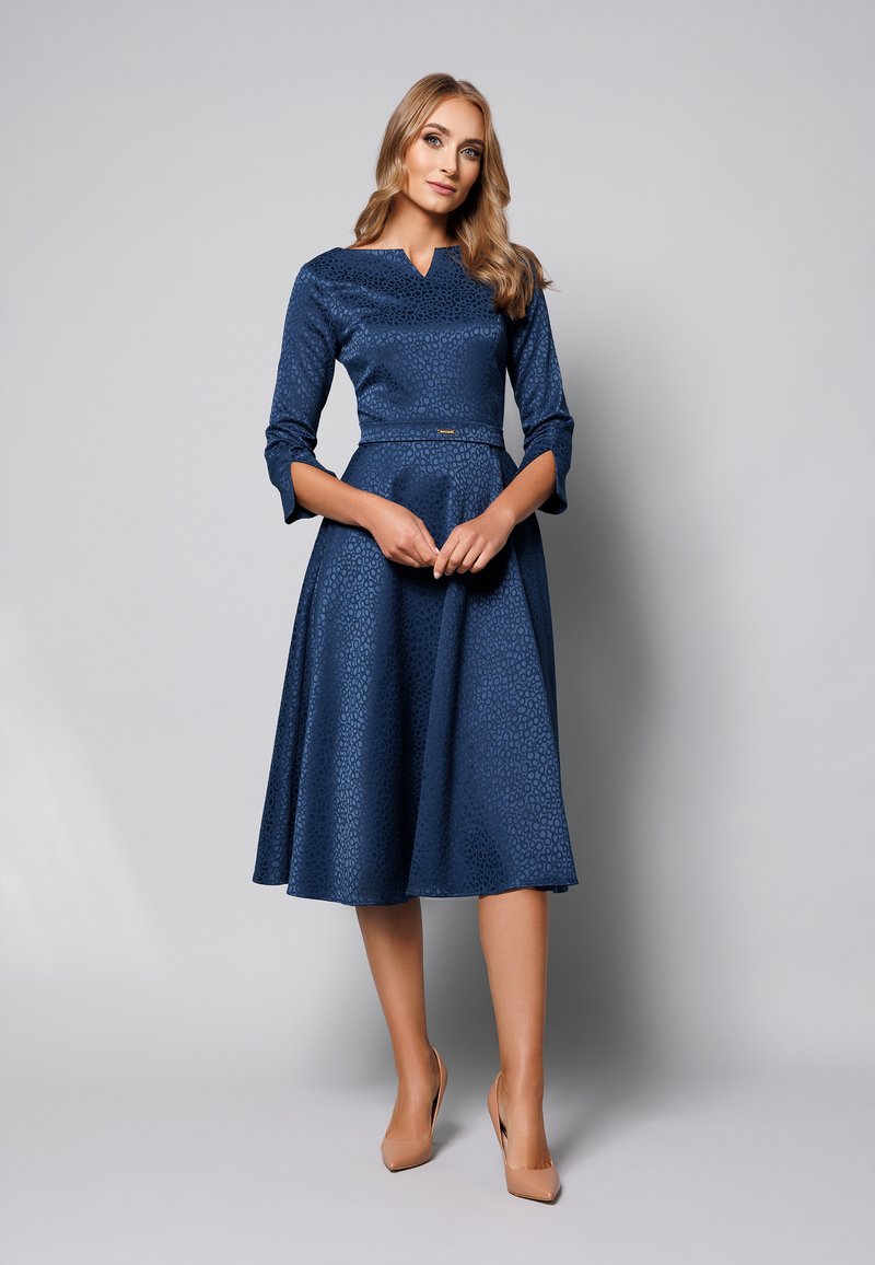 Granatowa sukienka z dopasowaną górą i rozkloszowanym dołem, z teksturowanym wzorem w kropki i rękawami do łokcia. Gładki materiał, subtelny pasek w talii.