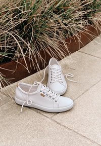 Zapatillas deportivas blancas con cordones colocadas sobre suelo de baldosas junto a un borde de jardín color óxido con hierba ornamental seca marrón y verde.